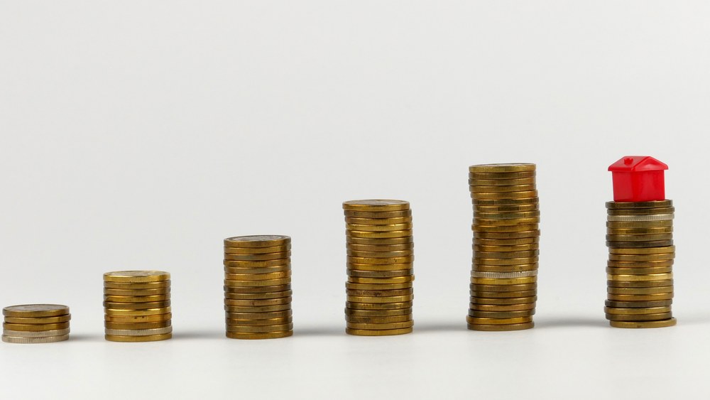 Stacks of coins growing taller from left to right with a small red house on the last stack
