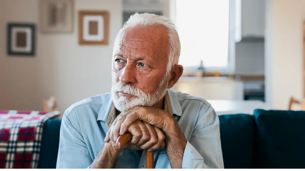 A senior man sitting on his sofa at home, resting his hands on a cane
