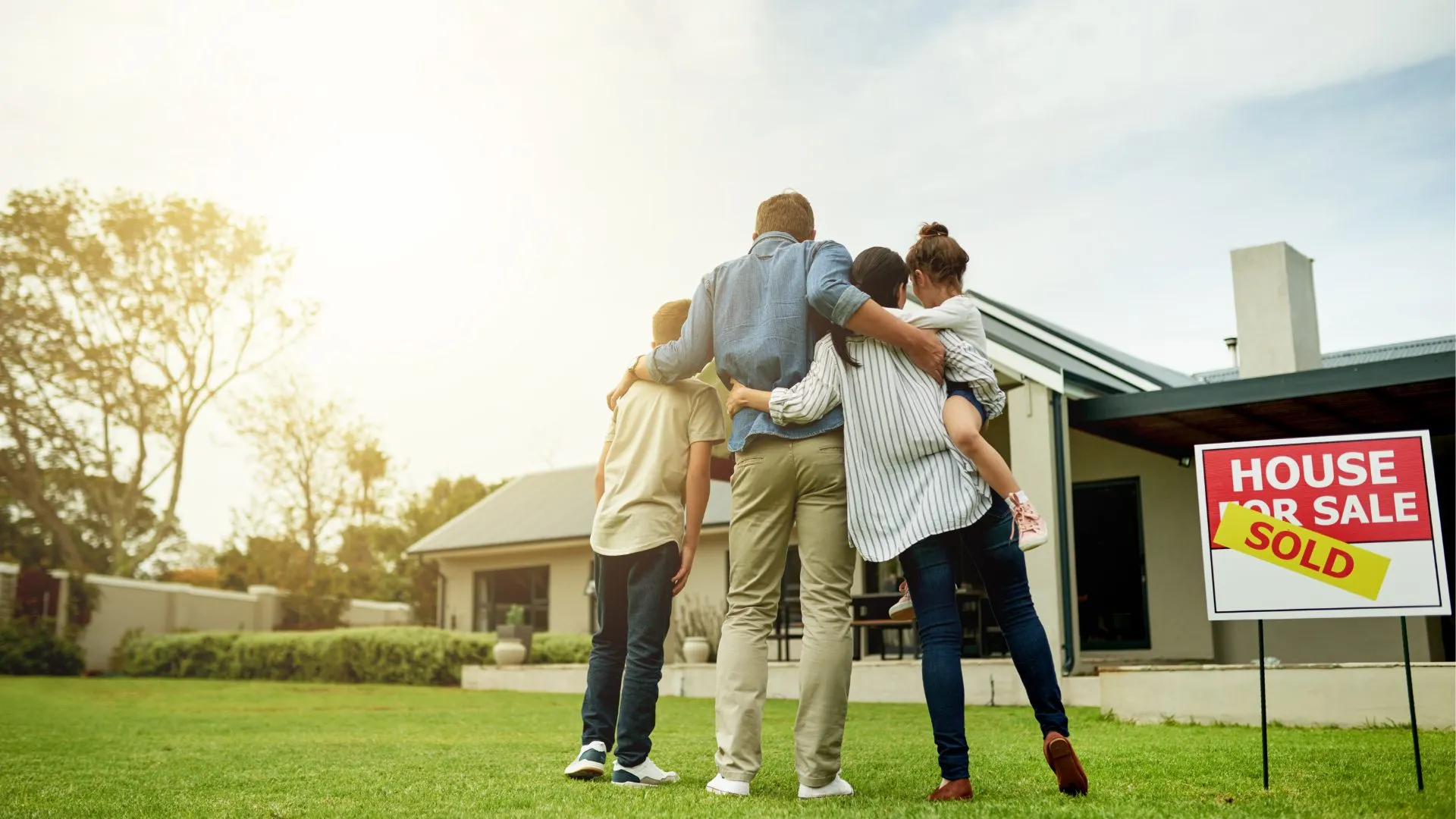 Family with their arms around each other looking at their new home beside a Sold sign