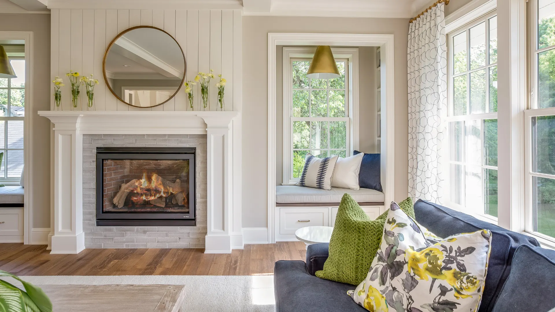 Bright, cozy living room with a white fireplace mantel, round mirror, window seat, and large windows