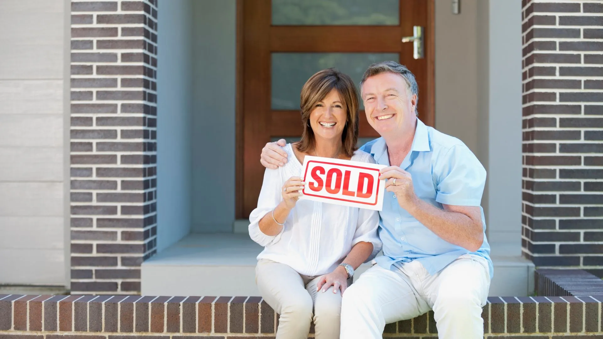 Smiling couple sitting on the front steps of their home holding a Sold sign